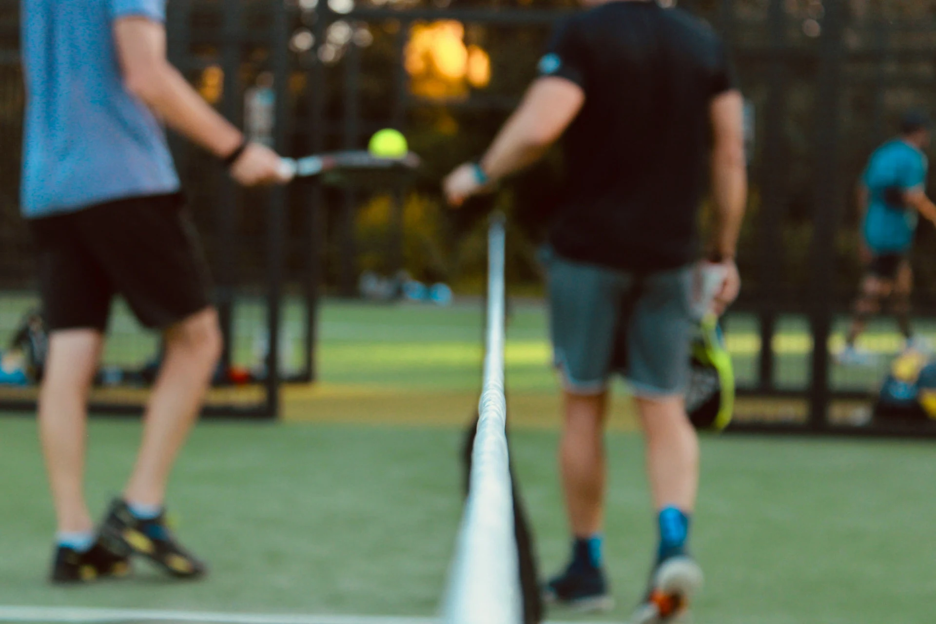 Jogadores de padel em quadra ao entardecer, com a rede em foco, uma bola no ar e raquetes na mão